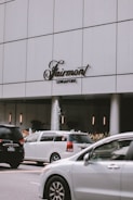 A modern city scene featuring a large white building with the Fairmont Singapore sign. Several cars are in motion on the road in front of the building. A small fountain is visible near the entrance, surrounded by plants.
