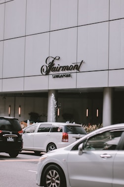 A modern city scene featuring a large white building with the Fairmont Singapore sign. Several cars are in motion on the road in front of the building. A small fountain is visible near the entrance, surrounded by plants.