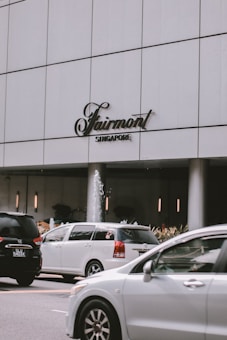 A modern city scene featuring a large white building with the Fairmont Singapore sign. Several cars are in motion on the road in front of the building. A small fountain is visible near the entrance, surrounded by plants.
