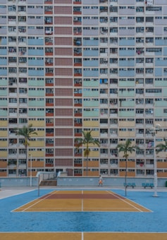 A tall residential building with a colorful facade featuring multiple rows of windows and balconies. In front of the building is a small sports court with a blue and yellow color scheme. Palm trees line the courtyard, and a lone person is walking near the court.