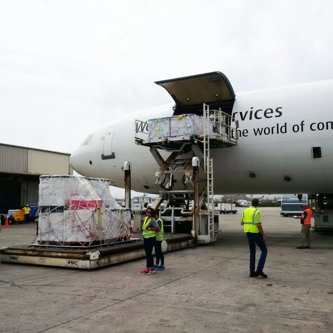 A cargo airplane is being loaded with large, wrapped packages at an airport. Several workers in high-visibility vests are present, overseeing the loading process. One worker is operating machinery to elevate the cargo into the airplane's open cargo hold. Various equipment and vehicles are situated around the plane.