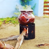 A traditional clay pot with geometric patterns is placed on a metal stove outdoors. Smoke rises from the pot, indicating it is being heated. Firewood is inserted into the stove, and part of a person's hand is visible, suggesting they are tending to the fire. The ground appears sandy, and there are hints of greenery and a blue wall in the background.