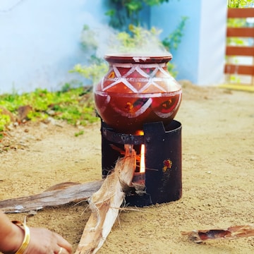 A traditional clay pot with geometric patterns is placed on a metal stove outdoors. Smoke rises from the pot, indicating it is being heated. Firewood is inserted into the stove, and part of a person's hand is visible, suggesting they are tending to the fire. The ground appears sandy, and there are hints of greenery and a blue wall in the background.