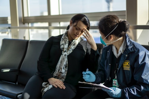 Two people are sitting in what appears to be an airport or waiting area. One person is wearing a scarf and appears to be unwell or stressed, touching their forehead. The other person is wearing a jacket with a CDC logo, gloves, and a face mask, and is holding a clipboard and pen, engaged in what seems to be a health screening or interview.