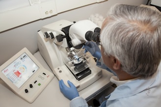 A person with gray hair is using a microscope in a laboratory setting. They are wearing blue gloves and focusing on a slide under the microscope. Next to them is an electronic display with controls, indicating sophisticated scientific equipment.