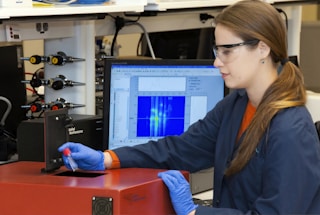 A person wearing safety glasses and gloves operates a scientific instrument, possibly in a laboratory setting. They hold a small vial near a device labeled 'Applied Nanofluorescence' while a computer monitor shows data graphs and charts in the background.