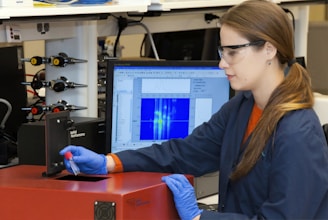 A person wearing safety glasses and gloves operates a scientific instrument, possibly in a laboratory setting. They hold a small vial near a device labeled 'Applied Nanofluorescence' while a computer monitor shows data graphs and charts in the background.
