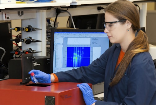 A person wearing safety glasses and gloves operates a scientific instrument, possibly in a laboratory setting. They hold a small vial near a device labeled 'Applied Nanofluorescence' while a computer monitor shows data graphs and charts in the background.