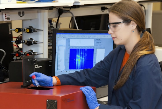 A person wearing safety glasses and gloves operates a scientific instrument, possibly in a laboratory setting. They hold a small vial near a device labeled 'Applied Nanofluorescence' while a computer monitor shows data graphs and charts in the background.