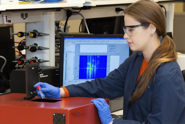 A person wearing safety glasses and gloves operates a scientific instrument, possibly in a laboratory setting. They hold a small vial near a device labeled 'Applied Nanofluorescence' while a computer monitor shows data graphs and charts in the background.