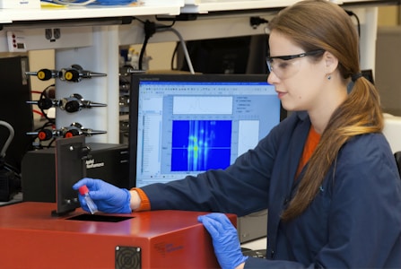 A person wearing safety glasses and gloves operates a scientific instrument, possibly in a laboratory setting. They hold a small vial near a device labeled 'Applied Nanofluorescence' while a computer monitor shows data graphs and charts in the background.