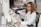 A woman wearing safety goggles and a lab coat is operating a large microscope in a laboratory setting. She is focused on the controls of the equipment. In the background, another person is working at a computer. The workspace is equipped with various scientific instruments and a keyboard.