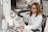 A woman wearing safety goggles and a lab coat is operating a large microscope in a laboratory setting. She is focused on the controls of the equipment. In the background, another person is working at a computer. The workspace is equipped with various scientific instruments and a keyboard.