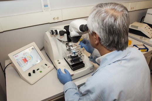 A person wearing a lab coat and blue gloves is looking through a microscope in a laboratory setting. There is a computer screen displaying data next to the microscope, and various laboratory equipment is present on the table.