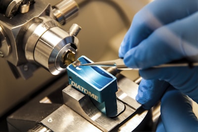 A close-up view of a precision laboratory instrument with a metal component and a blue holder labeled 'DIATOME'. A gloved hand is using tweezers to manipulate a small object, indicating a meticulous scientific or technical procedure.