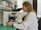 A scientist in a white lab coat is looking through a microscope in a laboratory setting. The background includes shelves filled with various laboratory equipment and supplies, such as bottles and containers. The laboratory has a clean and organized appearance.