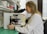 A scientist in a white lab coat is looking through a microscope in a laboratory setting. The background includes shelves filled with various laboratory equipment and supplies, such as bottles and containers. The laboratory has a clean and organized appearance.