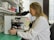 A scientist in a white lab coat is looking through a microscope in a laboratory setting. The background includes shelves filled with various laboratory equipment and supplies, such as bottles and containers. The laboratory has a clean and organized appearance.