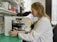 A scientist in a white lab coat is looking through a microscope in a laboratory setting. The background includes shelves filled with various laboratory equipment and supplies, such as bottles and containers. The laboratory has a clean and organized appearance.