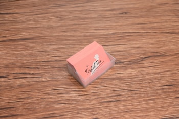 A stack of peach-colored business cards is placed inside a transparent holder on a wooden surface. The cards have a logo with the text 'The Twisted Trio' and a small silhouette of a head profile.