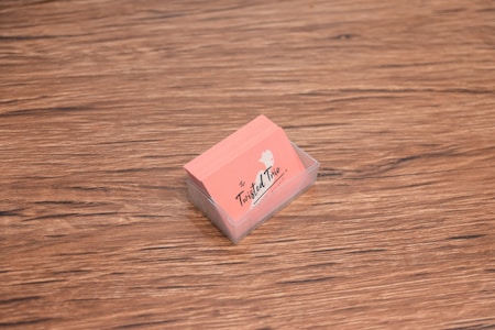 A stack of peach-colored business cards is placed inside a transparent holder on a wooden surface. The cards have a logo with the text 'The Twisted Trio' and a small silhouette of a head profile.