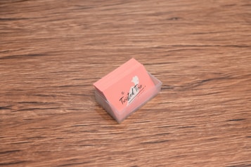 A stack of peach-colored business cards is placed inside a transparent holder on a wooden surface. The cards have a logo with the text 'The Twisted Trio' and a small silhouette of a head profile.