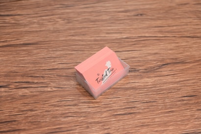 A stack of peach-colored business cards is placed inside a transparent holder on a wooden surface. The cards have a logo with the text 'The Twisted Trio' and a small silhouette of a head profile.