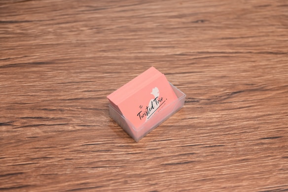 A stack of peach-colored business cards is placed inside a transparent holder on a wooden surface. The cards have a logo with the text 'The Twisted Trio' and a small silhouette of a head profile.