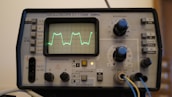 An oscilloscope displaying a waveform on its screen, surrounded by various knobs and buttons used for adjusting settings. The device appears to be an older model with labels in Russian, indicating voltage and other measurement settings. There are cables connected to inputs at the bottom.