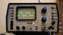 An oscilloscope displaying a waveform on its screen, surrounded by various knobs and buttons used for adjusting settings. The device appears to be an older model with labels in Russian, indicating voltage and other measurement settings. There are cables connected to inputs at the bottom.