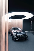 A sleek black luxury car is displayed in an elegant showroom with a modern and minimalist design. The car is surrounded by vertical strands of light brown string, which add an artistic touch. Above the vehicle, a large circular light fixture provides illumination, casting a soft white glow on the scene.