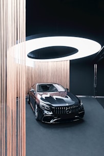 A sleek black luxury car is displayed in an elegant showroom with a modern and minimalist design. The car is surrounded by vertical strands of light brown string, which add an artistic touch. Above the vehicle, a large circular light fixture provides illumination, casting a soft white glow on the scene.