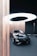 A sleek black luxury car is displayed in an elegant showroom with a modern and minimalist design. The car is surrounded by vertical strands of light brown string, which add an artistic touch. Above the vehicle, a large circular light fixture provides illumination, casting a soft white glow on the scene.