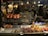 A street food stall is filled with a variety of grilled foods, including whole chickens, fish, and skewers of meat and vegetables. The food is displayed on metal trays and cooking racks. The background shows a bustling night market with people moving around, illuminated by bright lights hanging from above.