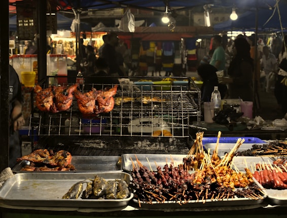 A street food stall is filled with a variety of grilled foods, including whole chickens, fish, and skewers of meat and vegetables. The food is displayed on metal trays and cooking racks. The background shows a bustling night market with people moving around, illuminated by bright lights hanging from above.