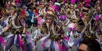 A group of performers are participating in a parade. They are wearing elaborate and colorful costumes with intricate designs, featuring purple, pink, and gold accents. Their outfits include feathered headdresses, sequined fabric, and various adornments. The performers are marching energetically through a street lined with spectators.