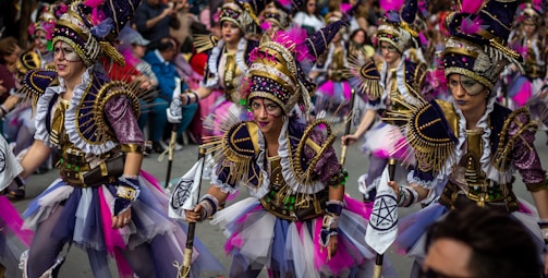 A group of performers are participating in a parade. They are wearing elaborate and colorful costumes with intricate designs, featuring purple, pink, and gold accents. Their outfits include feathered headdresses, sequined fabric, and various adornments. The performers are marching energetically through a street lined with spectators.