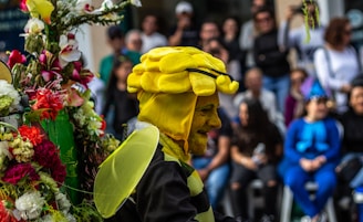 A person is dressed in a vibrant bee costume with yellow and black stripes, including a headpiece shaped like a bee's head and a pair of wings. They are standing next to a colorful array of flowers. In the background, a crowd of onlookers is gathered, with some dressed in costumes, indicating a festive or parade-like atmosphere.
