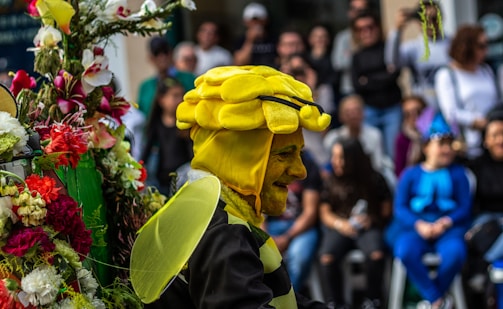 A person is dressed in a vibrant bee costume with yellow and black stripes, including a headpiece shaped like a bee's head and a pair of wings. They are standing next to a colorful array of flowers. In the background, a crowd of onlookers is gathered, with some dressed in costumes, indicating a festive or parade-like atmosphere.