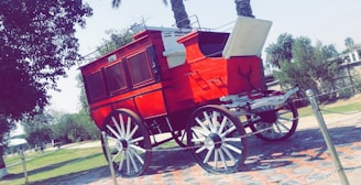 A vividly painted red stagecoach with white wheels, parked on a paved surface surrounded by a grassy area and palm trees. The coach is enclosed with a rear door open, suggesting a vintage or historical theme.