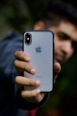 A person holds an iPhone with a silver back and black edges in one hand. The focus is on the smartphone, displaying the Apple logo and the word 'iPhone' with clarity. The background is blurred, obscuring the person's face and any details behind.