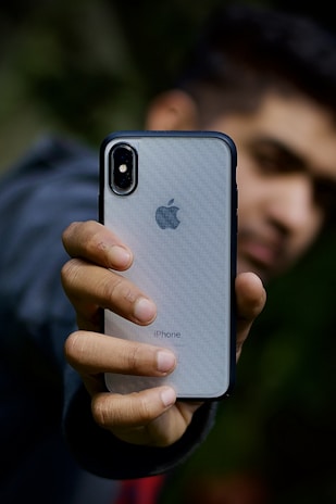 A person holds an iPhone with a silver back and black edges in one hand. The focus is on the smartphone, displaying the Apple logo and the word 'iPhone' with clarity. The background is blurred, obscuring the person's face and any details behind.