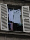 A window with white shutters is open, revealing clothes hanging to dry. The garments include shirts and towels in various shades of blue, white, and dark colors. The window is part of a gray building, and the clothes are draped over a railing to air out.