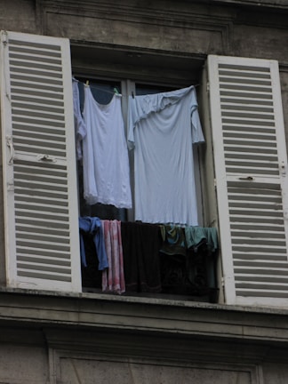 A window with white shutters is open, revealing clothes hanging to dry. The garments include shirts and towels in various shades of blue, white, and dark colors. The window is part of a gray building, and the clothes are draped over a railing to air out.