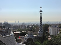 A cityscape view featuring a mosaic-covered building in the foreground with a spiraling tower topped by a cross. The background displays a sprawling urban skyline under a clear blue sky. The architecture exhibits colorful and intricate designs characteristic of Catalan modernism.