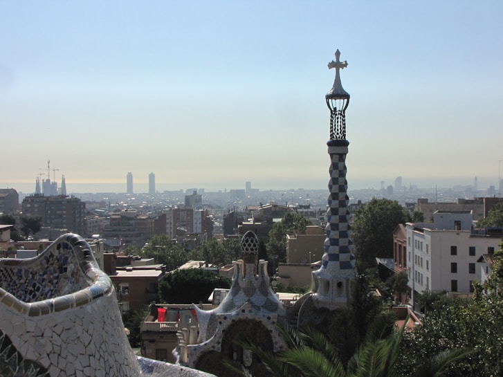 A cityscape view featuring a mosaic-covered building in the foreground with a spiraling tower topped by a cross. The background displays a sprawling urban skyline under a clear blue sky. The architecture exhibits colorful and intricate designs characteristic of Catalan modernism.