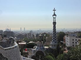 A cityscape view featuring a mosaic-covered building in the foreground with a spiraling tower topped by a cross. The background displays a sprawling urban skyline under a clear blue sky. The architecture exhibits colorful and intricate designs characteristic of Catalan modernism.