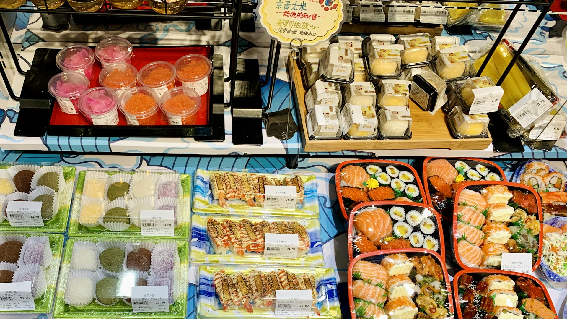 A variety of packaged sushi and desserts displayed on shelves. The sushi includes salmon nigiri, rolls, and assortment platters, while the desserts seem to consist of colorful mochi and parfaits in cups. Labels with prices are visible on many items.
