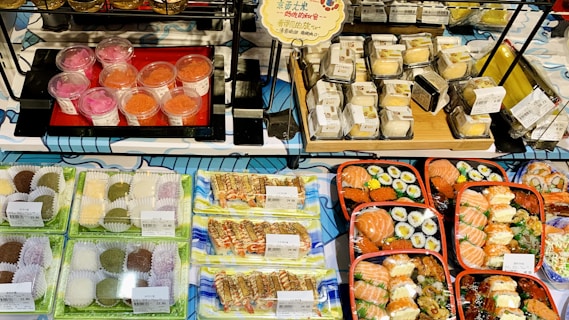 A variety of packaged sushi and desserts displayed on shelves. The sushi includes salmon nigiri, rolls, and assortment platters, while the desserts seem to consist of colorful mochi and parfaits in cups. Labels with prices are visible on many items.