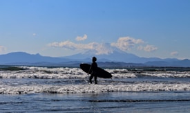 A person is walking along the shore with a surfboard under their arm, heading towards the ocean. Waves are gently crashing at their feet, and a majestic snow-capped mountain looms in the background under a clear blue sky. The scene captures a sense of adventure and serenity.